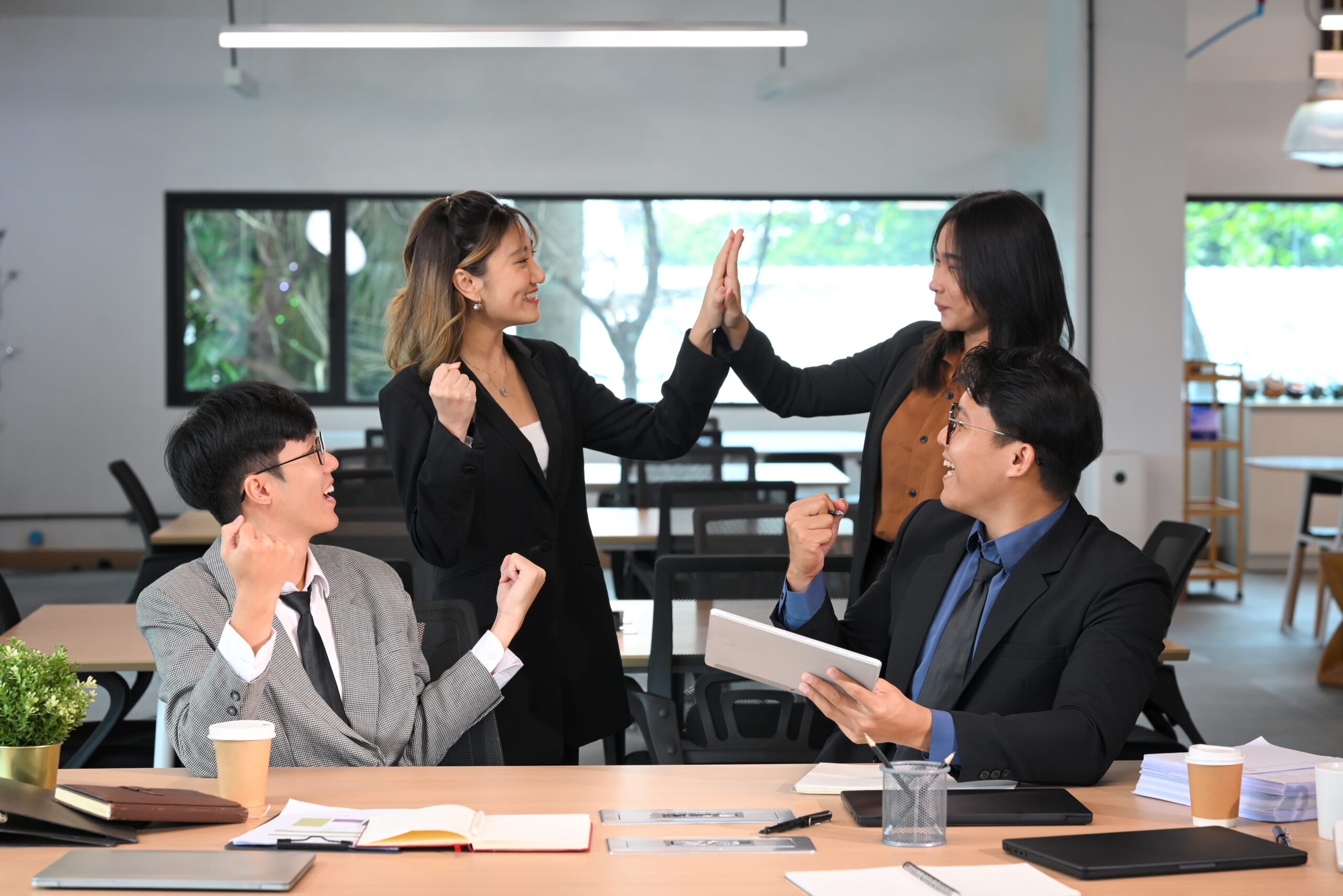Four colleagues in business attire celebrate and high-five in a modern office.