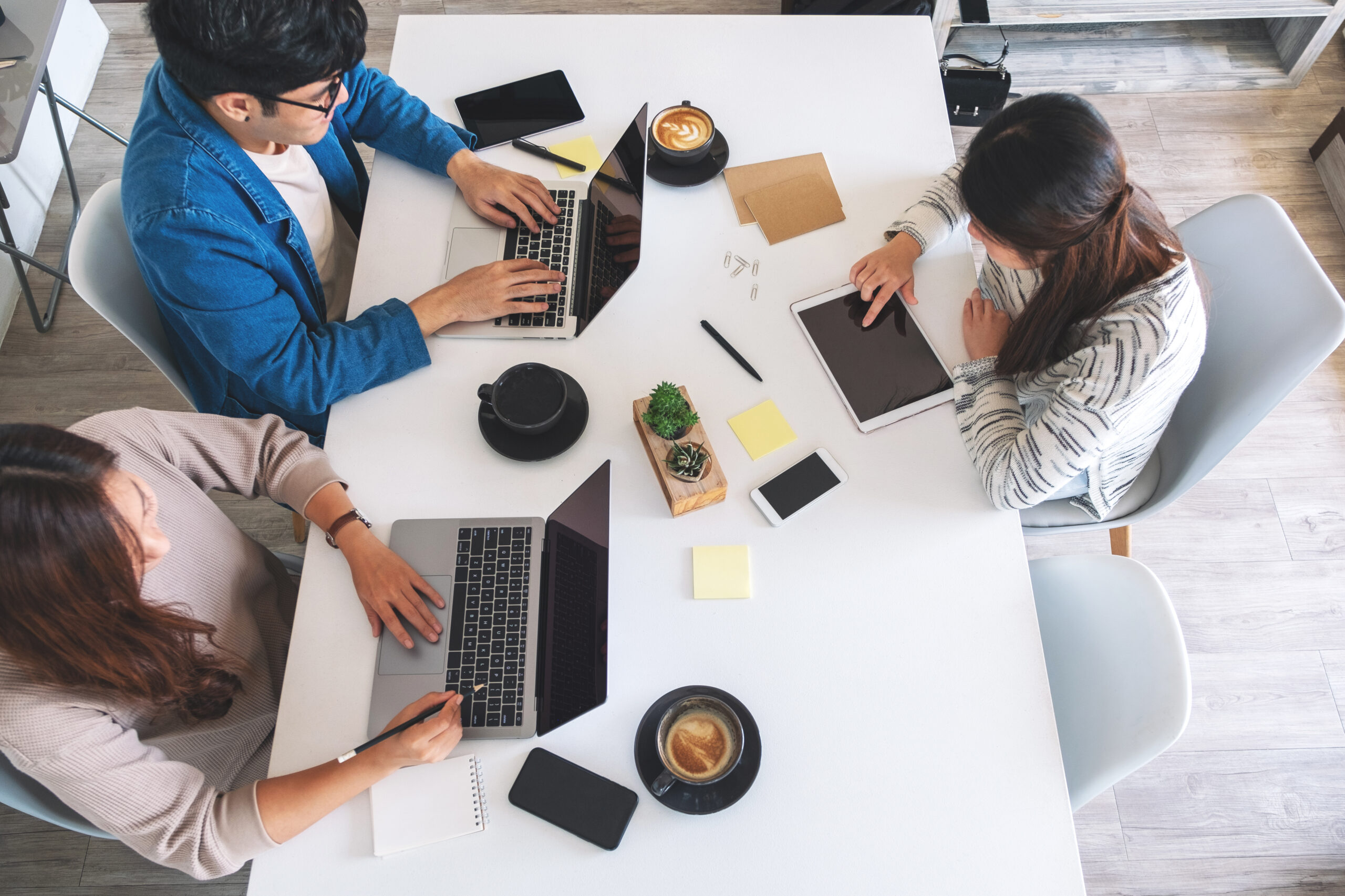 Three people working at a table with laptops, tablet, notebooks, and coffee.