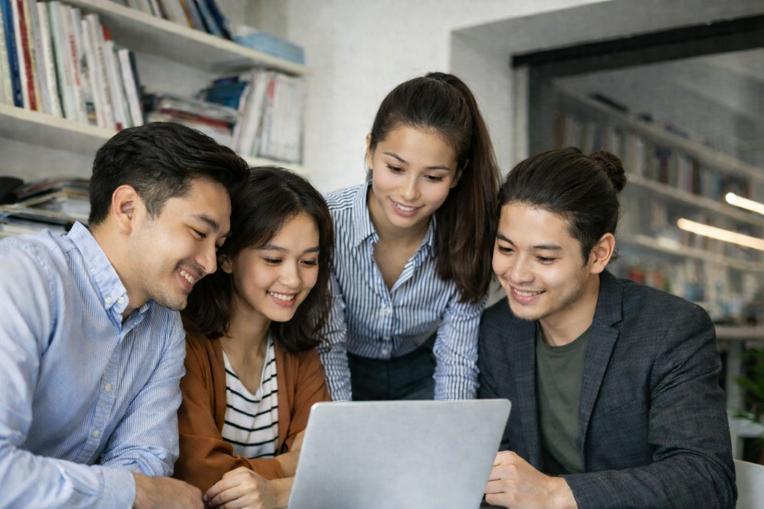 Four young adults smiling and looking at a laptop together in an office setting.